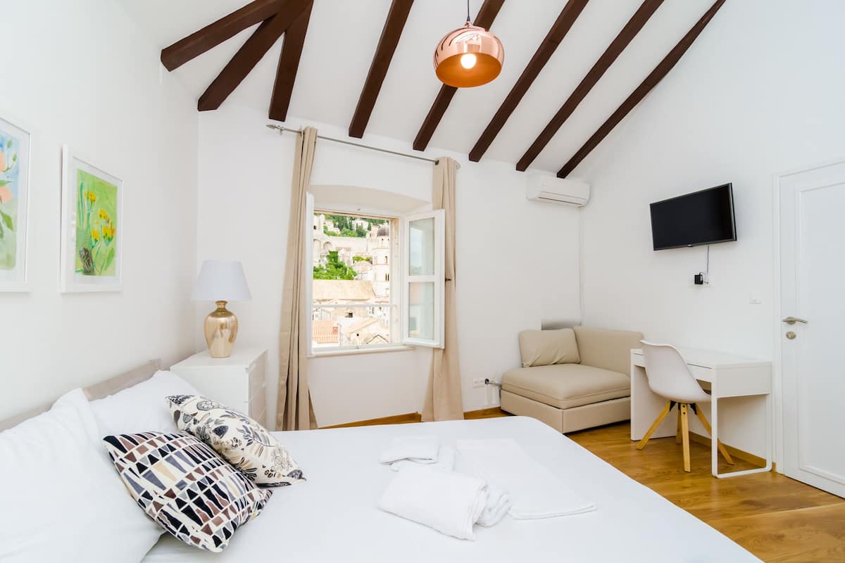 A bedroom with a bed, a desk, a chair, a TV, and a window with curtains.