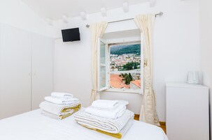 Quarto de hotel com uma cama, televisão na parede, janela com cortinas e vista para a cidade.