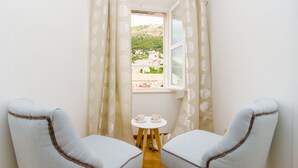 Una habitación pequeña con dos sillas mirando a una ventana, una mesa pequeña con tazas y vistas a una colina con edificios.