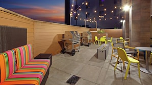 An outdoor patio area with a colorful striped sofa, yellow chairs, and barbecue grills. - Home2 Suites by Hilton Alameda Oakland Airport (Alameda)
