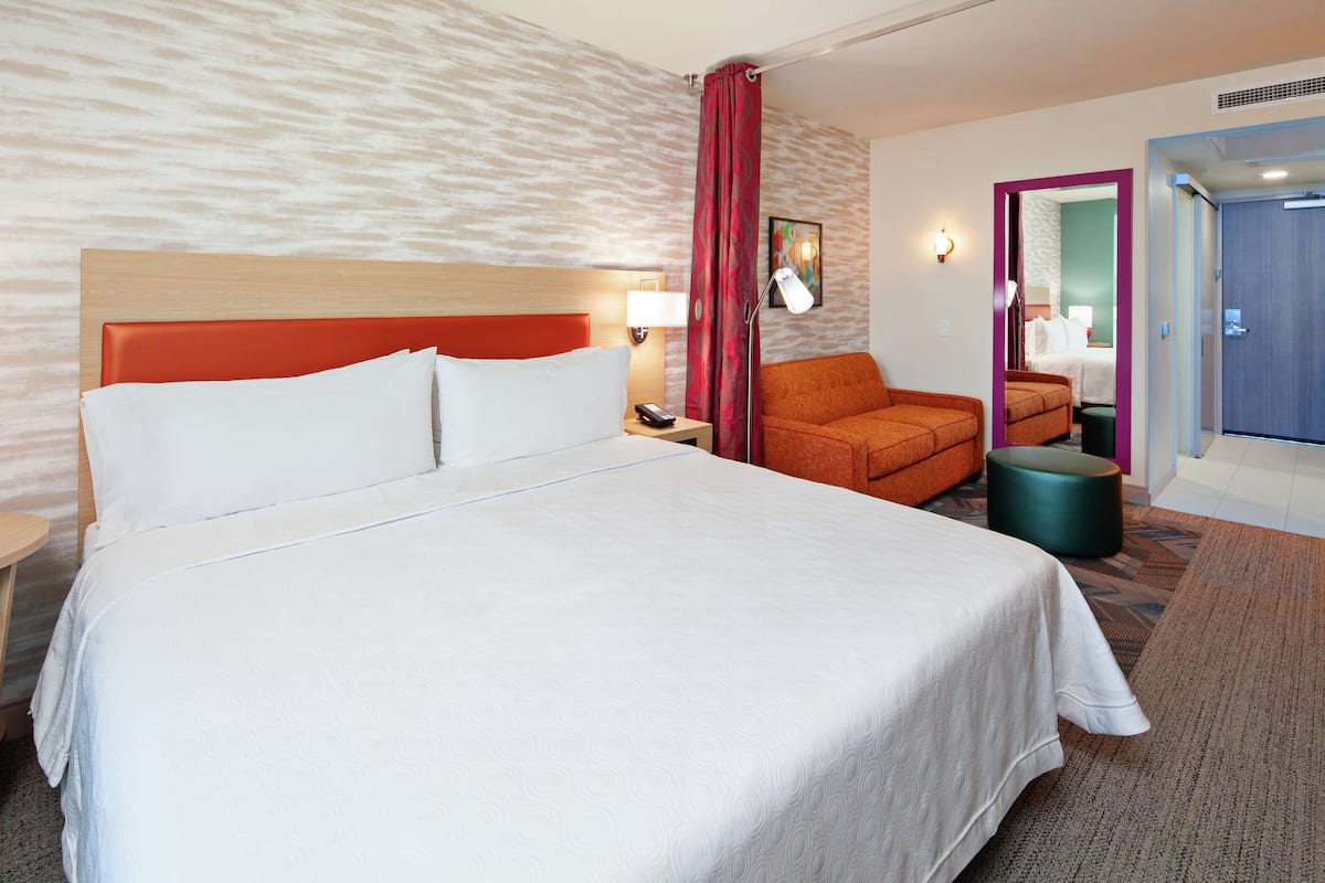 Hotellihuone, jossa on suuri sänky, työpöytä lampun kanssa, tuoli ja pieni pyöreä pöytä.