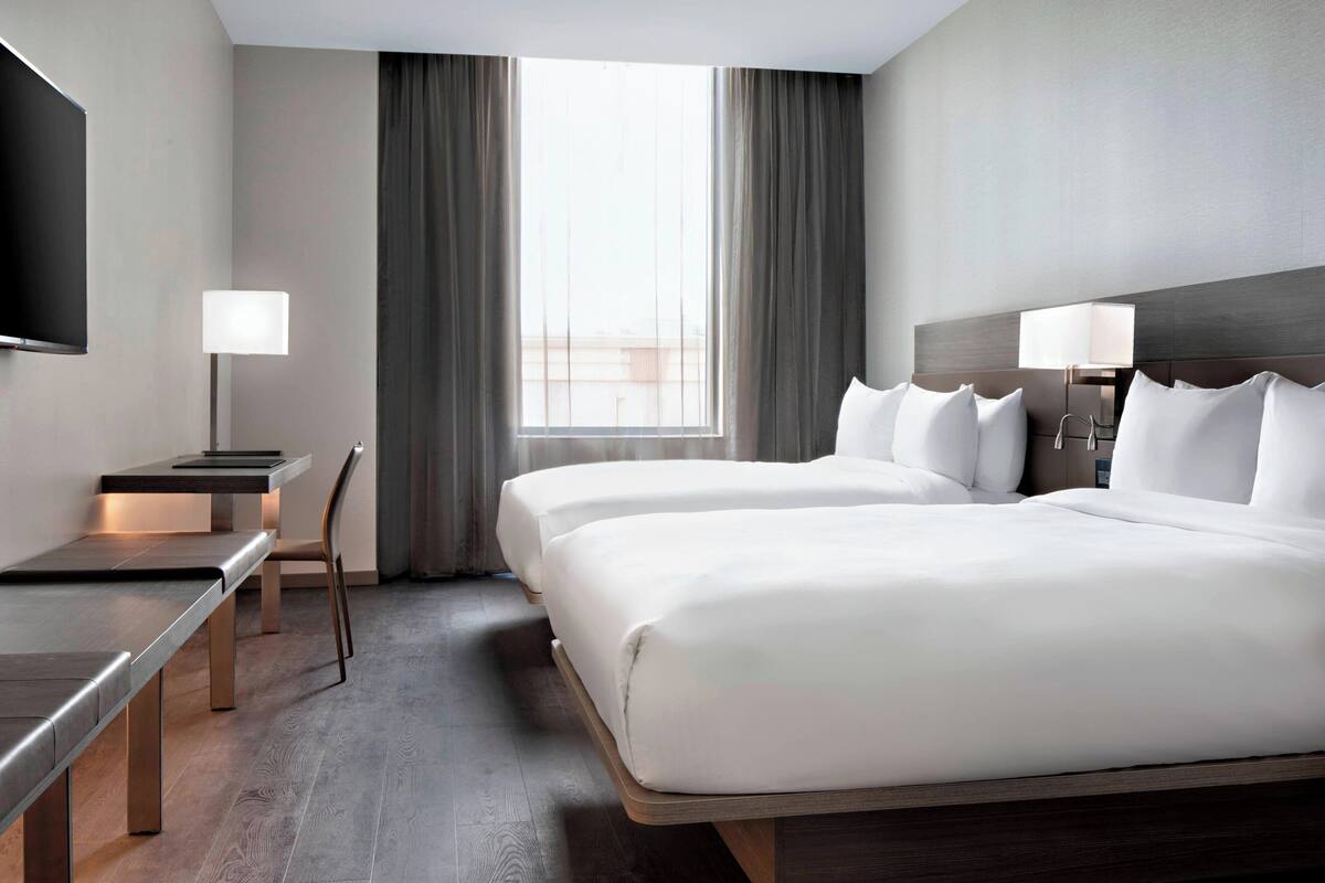 A hotel room with two beds, a desk, a chair, a TV, and a window with curtains.