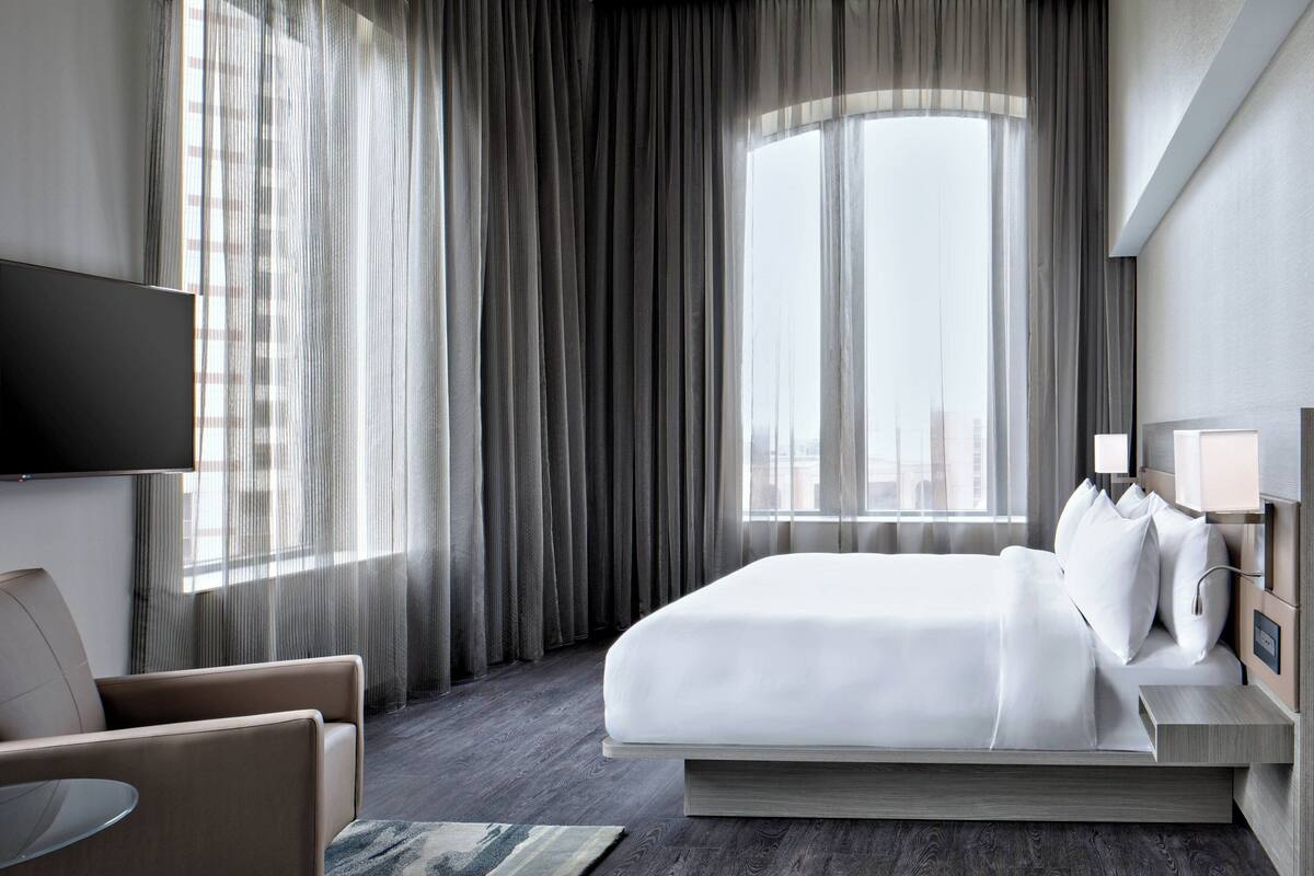 A modern hotel room with a large bed, a flat-screen TV, a bedside table with lamps, and a view of the city through sheer curtains.