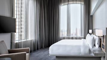 A modern hotel room with a large bed, a flat-screen TV, a bedside table with lamps, and a view of the city through sheer curtains.