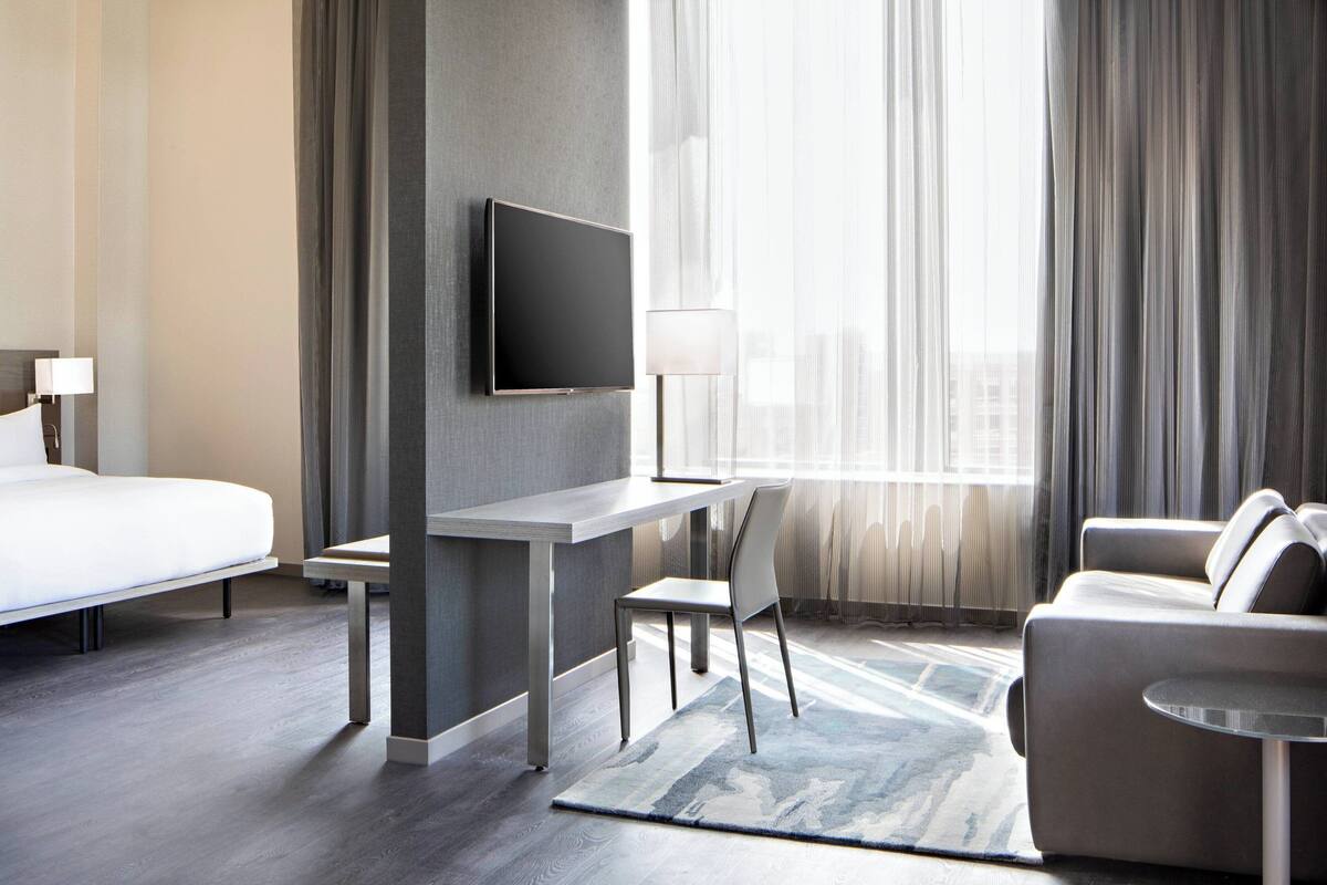 A modern hotel room with a bed, a desk, a chair, a sofa, and a TV mounted on the wall.