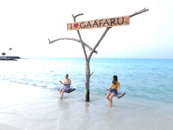 A beach scene with a wooden sign that reads "I ♥ GAAFARU" and two people enjoying the water.