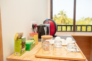 A tray with green tea boxes, a water bottle, a coffee maker, and glasses on a wooden table near a window with a view of palm trees.