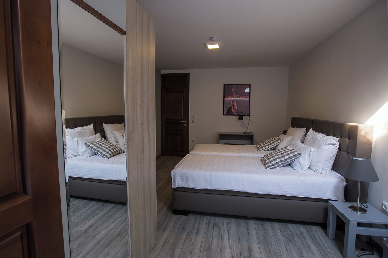 Ein Hotelzimmer mit zwei Betten, Holzboden, Spiegel, Tür, Nachttisch mit Lampe und einem an der Wand montierten Fernseher.