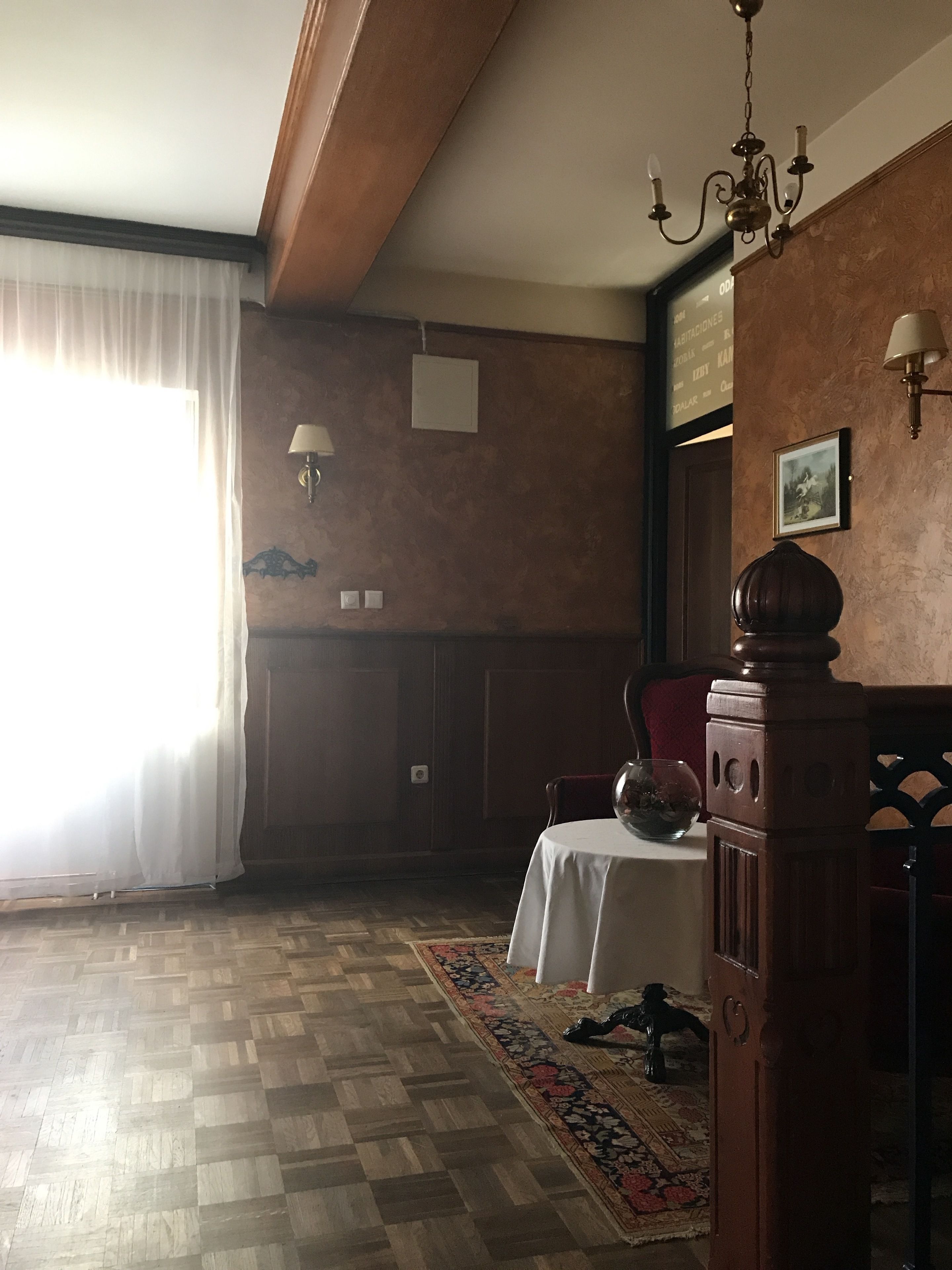 A room with a wooden floor, a dining table with chairs, a chandelier, and a large window with sheer curtains.