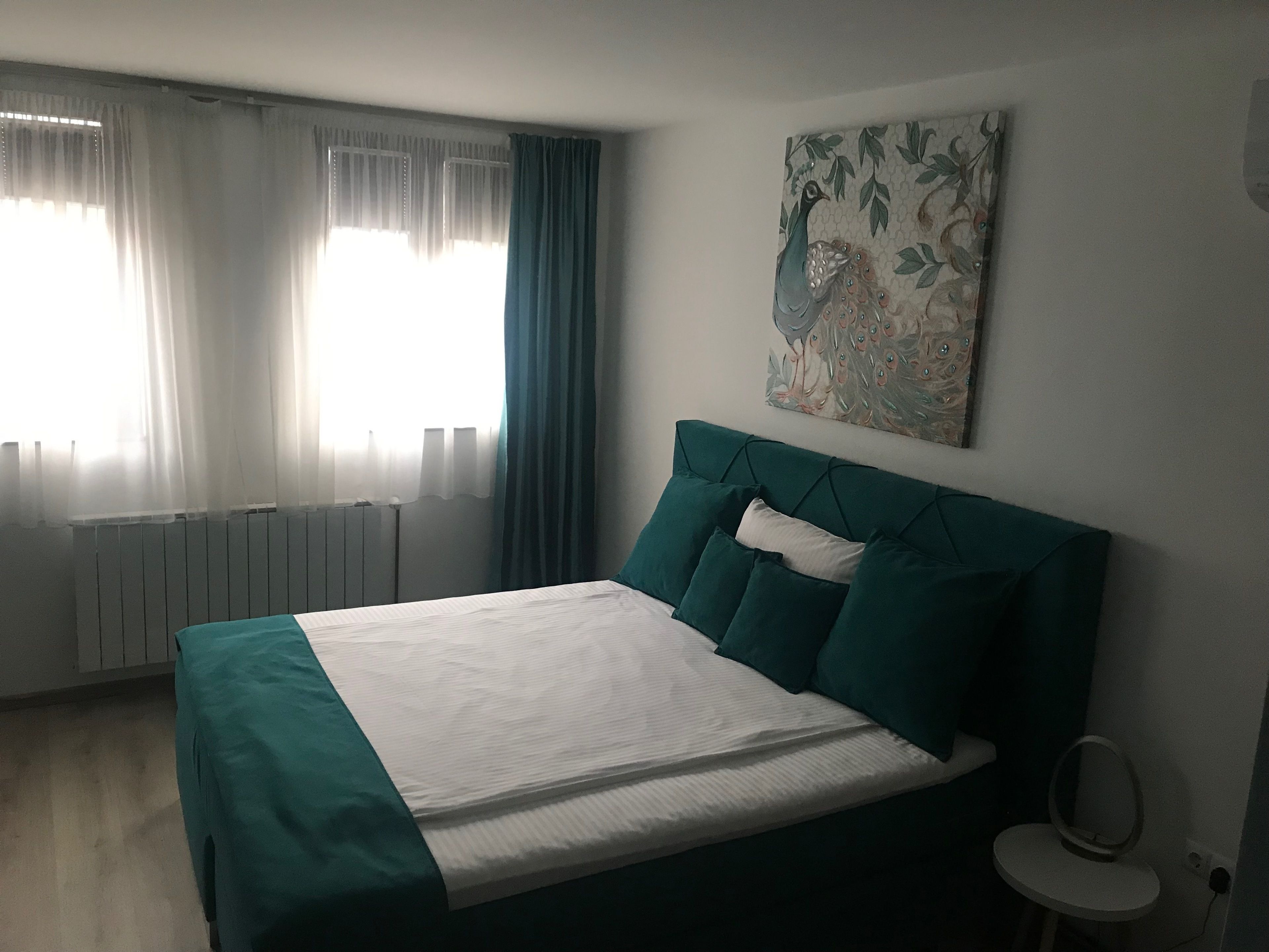 A bedroom with a bed, a radiator, a window with curtains, and a wall-mounted artwork.