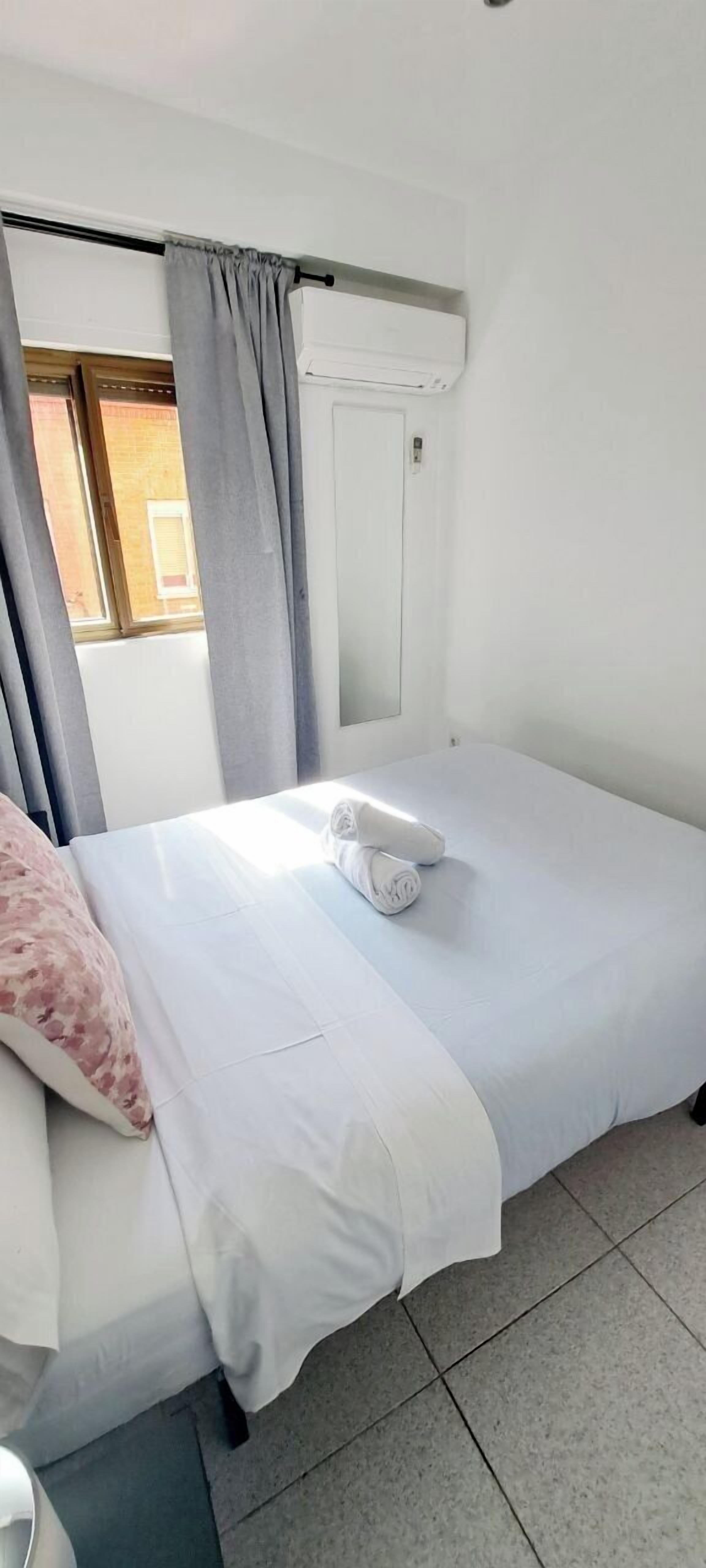 Un lit simple avec un matelas et des oreillers blancs, des rideaux gris et une fenêtre donnant sur un bâtiment en briques.
