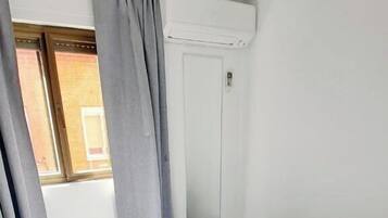 Un lit simple avec un matelas et des oreillers blancs, des rideaux gris et une fenêtre donnant sur un bâtiment en briques.
