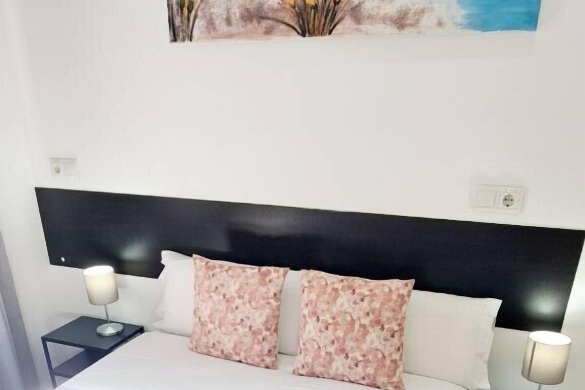 Una cama bien hecha con sábanas blancas y una toalla enrollada, un cabecero negro, dos mesitas de noche con lámparas y un cuadro con una escena costera sobre la cama.