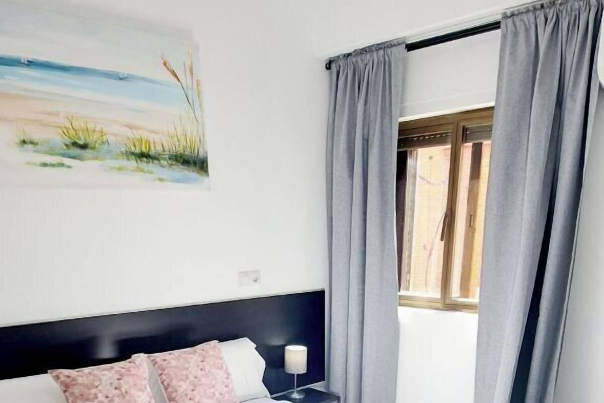 Una cama bien hecha con ropa blanca y una toalla enrollada, una mesita de noche con una lámpara, y un cuadro de una playa en la pared.