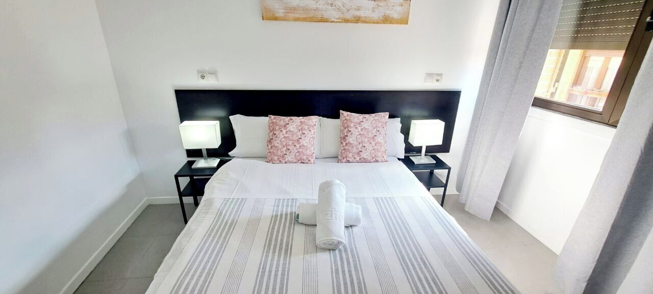 A hotel room with a bed, two bedside lamps, a headboard, and a window with curtains.