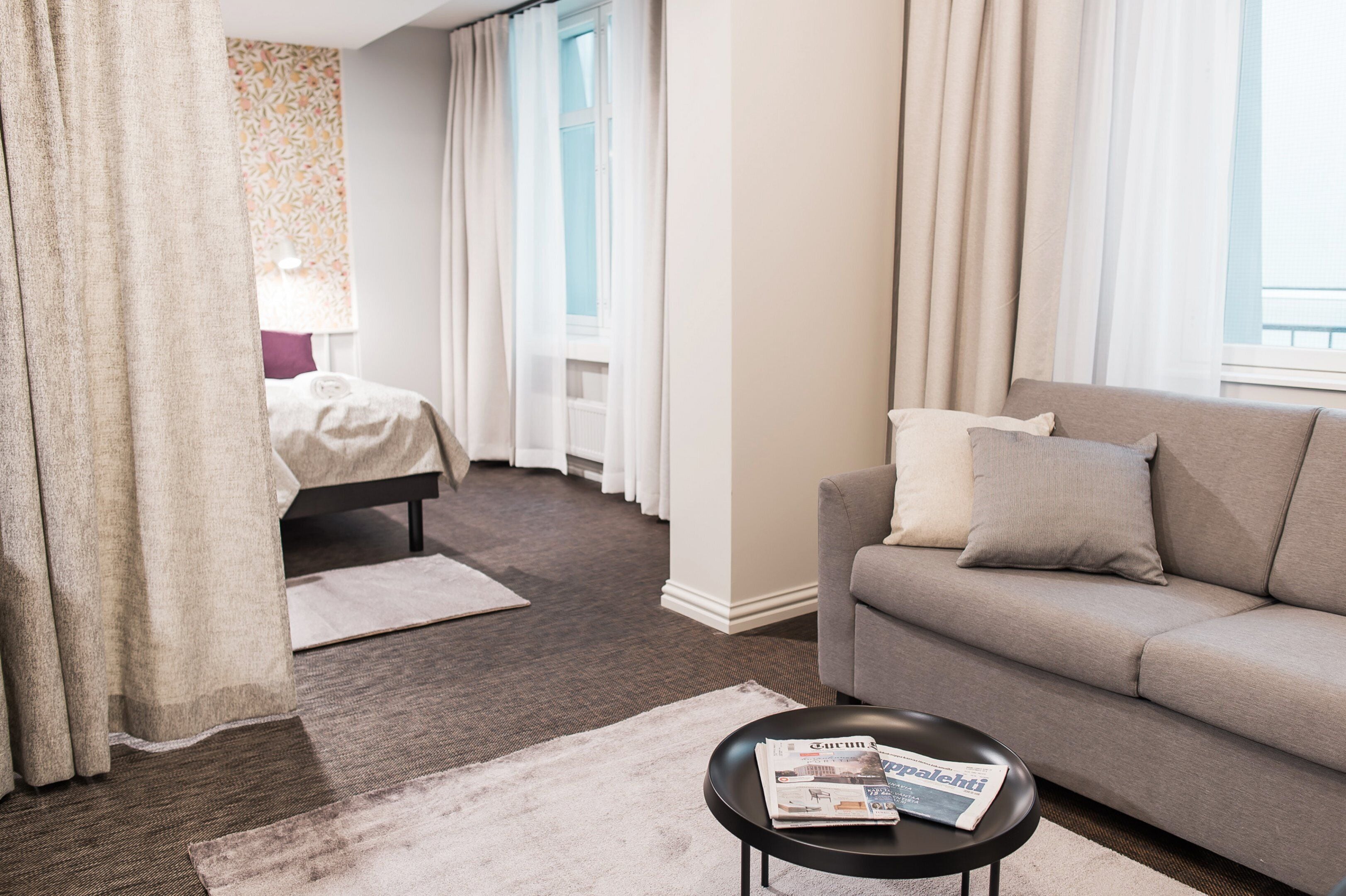 A hotel room with a bed, a sofa, a coffee table with a magazine, and a window with curtains.
