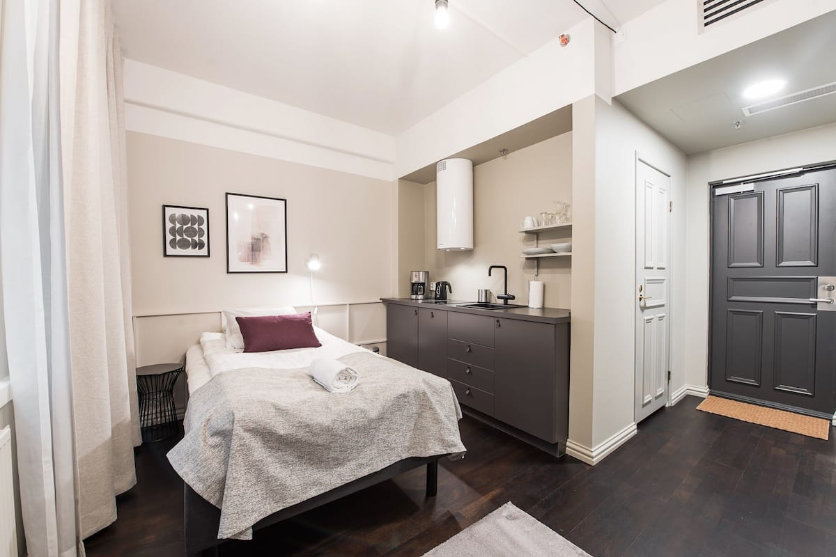 A modern bedroom with a bed, bedside table, and a small desk with a chair. There is a wardrobe and a door with a handle.