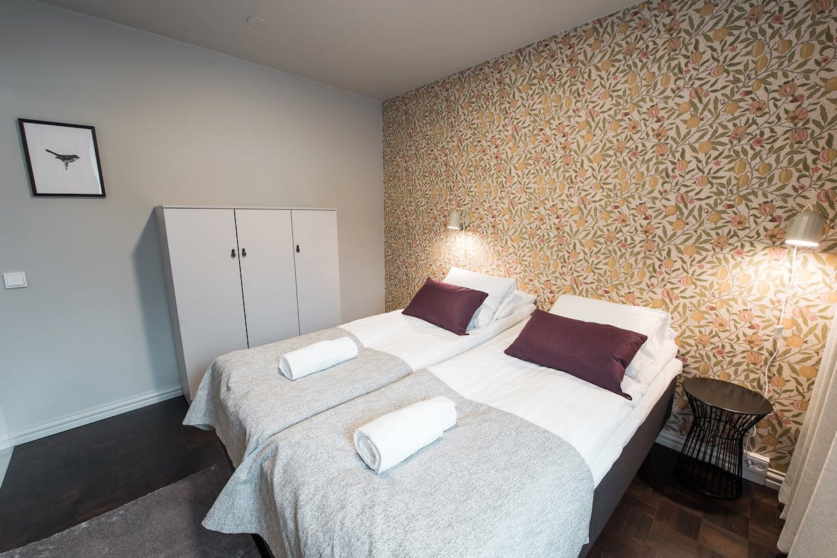 A hotel room with two beds, a wardrobe, a small table, and a wall with a floral pattern.