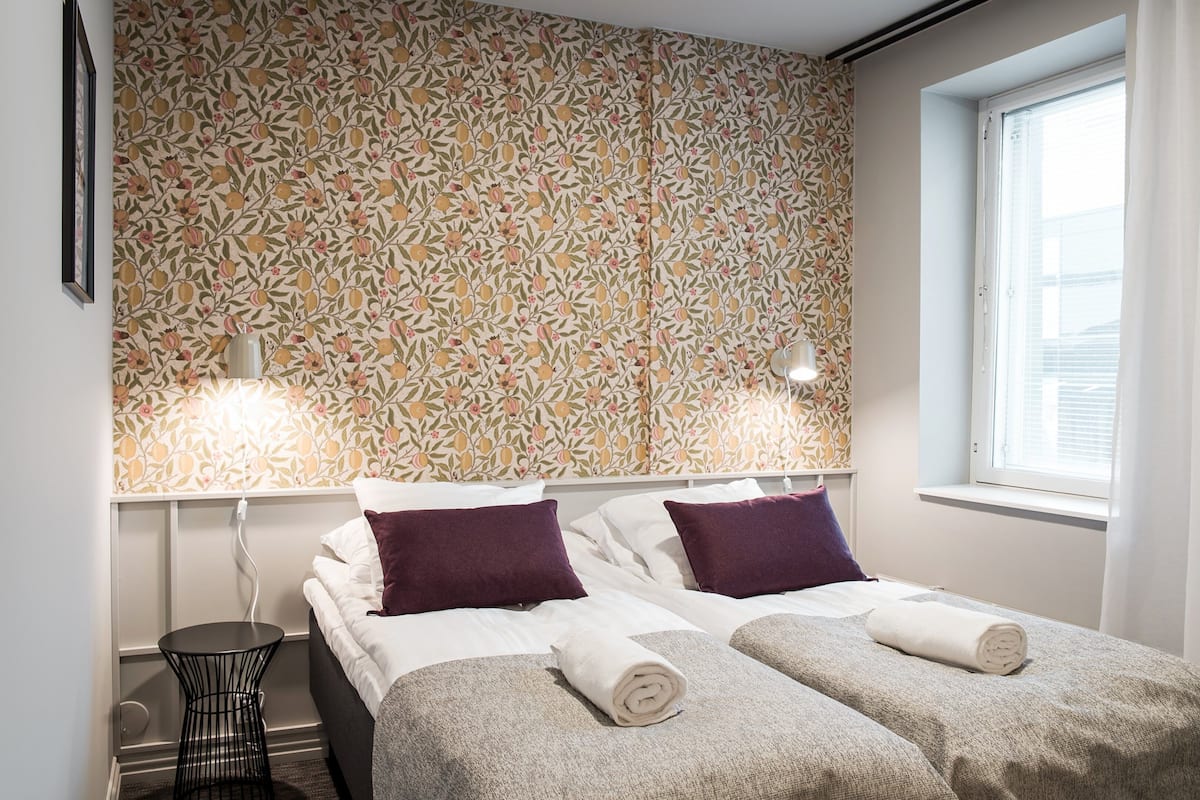 A double bed with two pillows, a small round table, and a wall with a floral pattern.