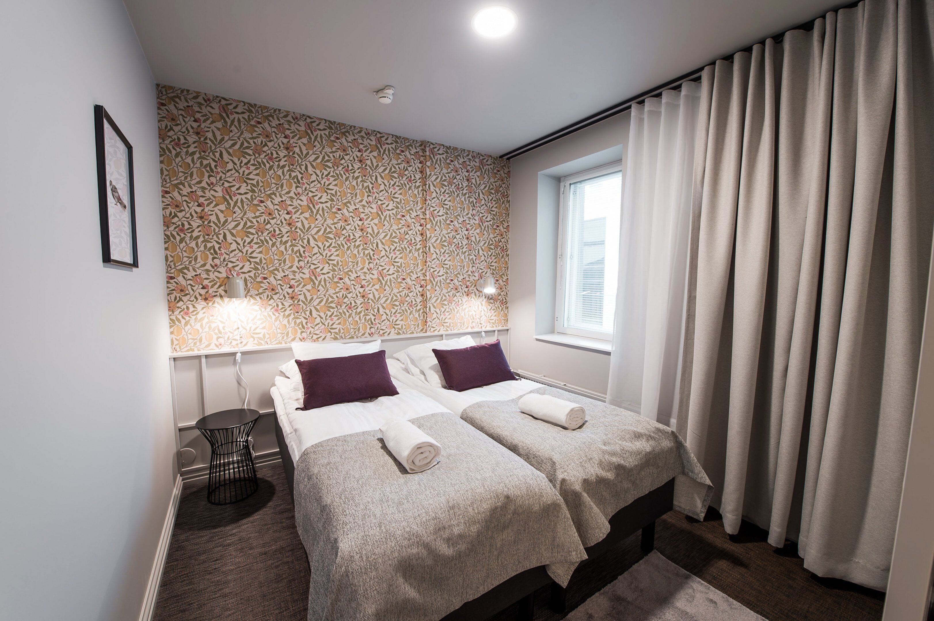 A hotel room with two beds, a patterned wallpaper, a small table, and a window with curtains.