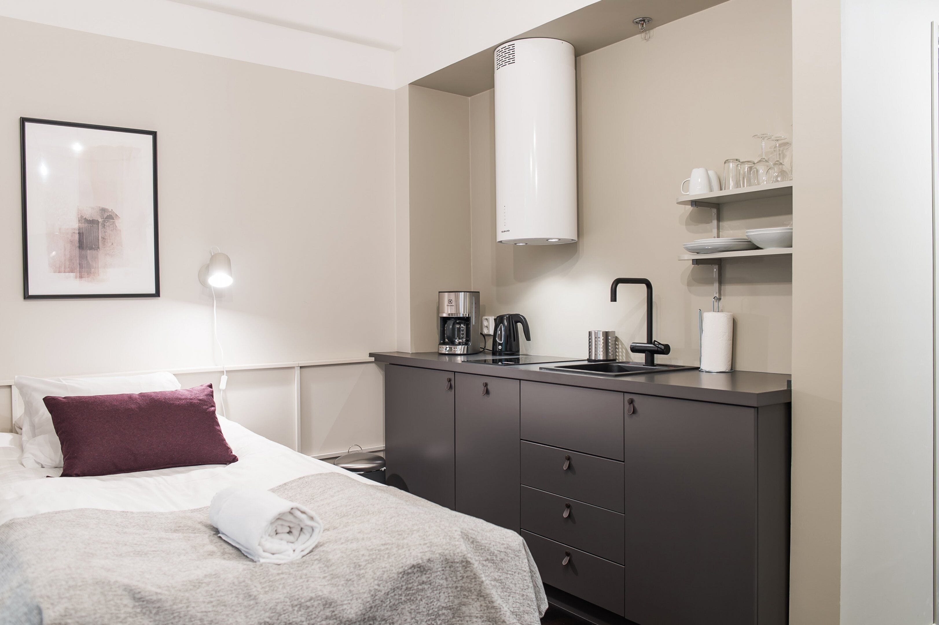 A modern hotel room with a bed, a bedside table with a lamp, a built-in kitchen with a coffee maker, and a large white vent.