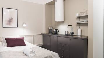 A modern hotel room with a bed, a bedside table with a lamp, a built-in kitchen with a coffee maker, and a large white vent.