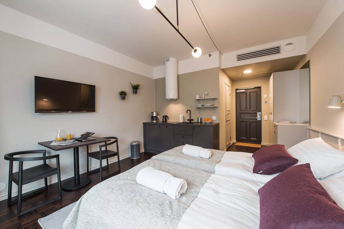 A modern hotel room with a bed, two bedside tables, a desk with chairs, a flat-screen TV, a kitchenette, and a bathroom.