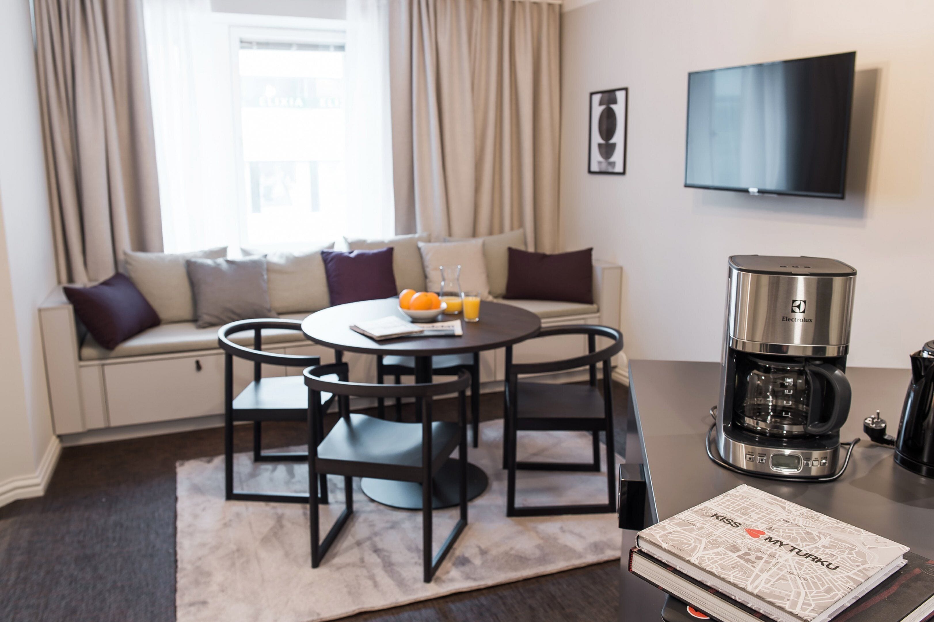 A modern living room with a sofa, a round table with chairs, a coffee maker, and a flat-screen TV.