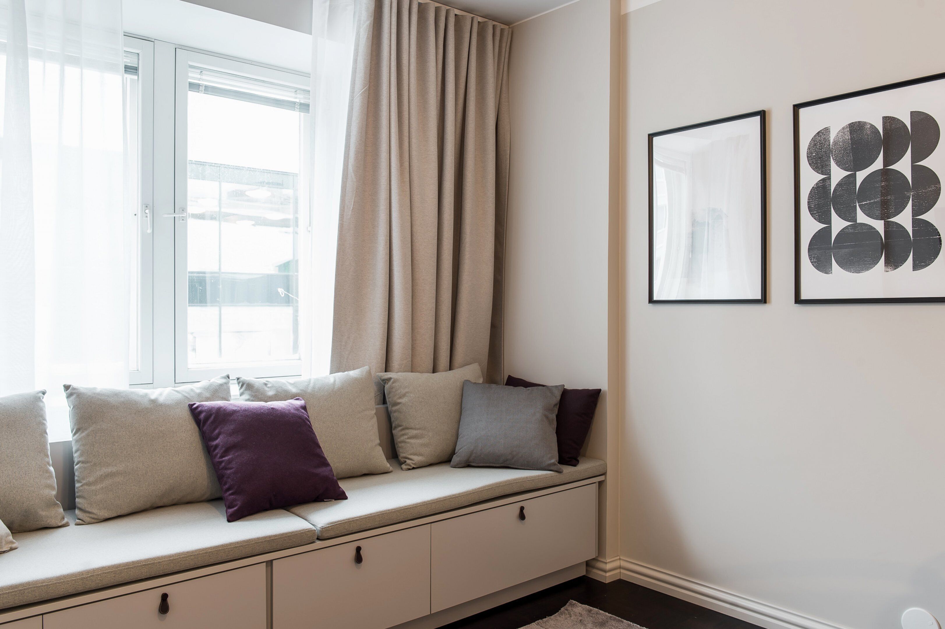 Une pièce avec une banquette intégrée dotée de coussins, une fenêtre avec des rideaux et deux œuvres d’art encadrées accrochées au mur.