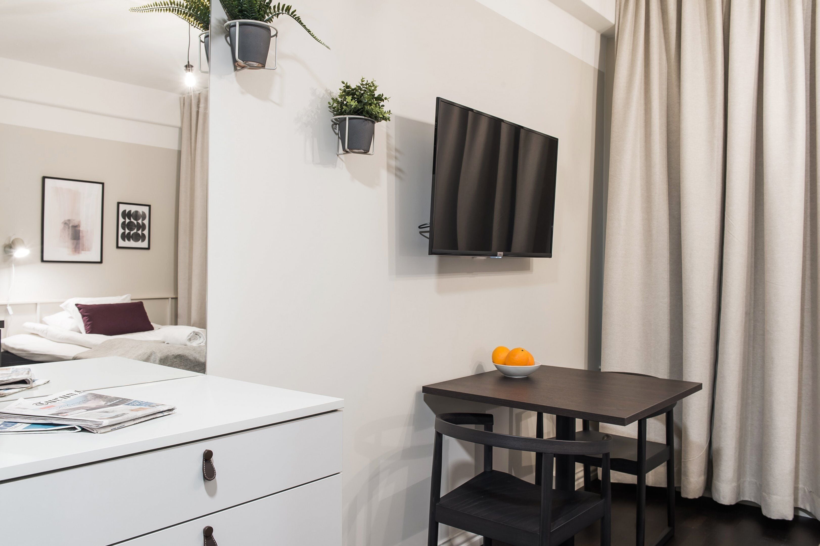 Une chambre d’hôtel moderne comprenant un lit, un bureau blanc avec des tiroirs, une petite table avec un saladier de fruits, un téléviseur fixé au mur et deux jardinières murales.