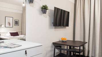 Une chambre d’hôtel moderne comprenant un lit, un bureau blanc avec des tiroirs, une petite table avec un saladier de fruits, un téléviseur fixé au mur et deux jardinières murales.