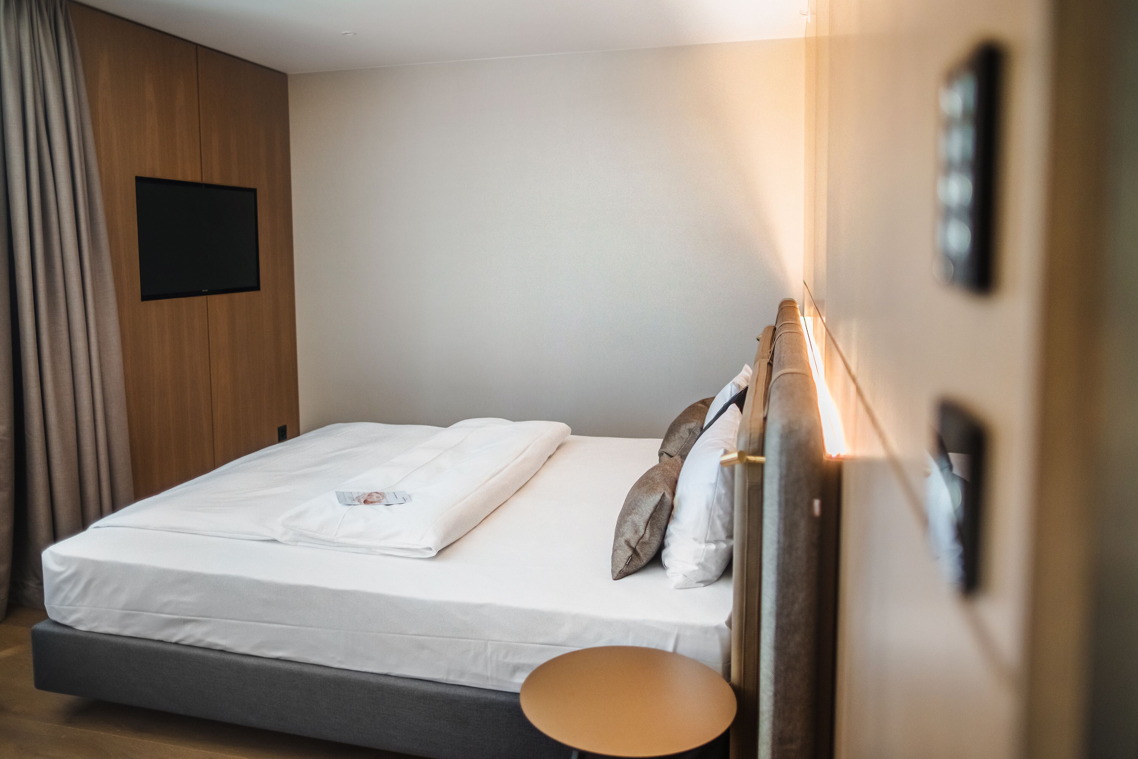Una cama bien hecha con colchón y almohadas blancos, una mesita redonda pequeña y un televisor montado en la pared.