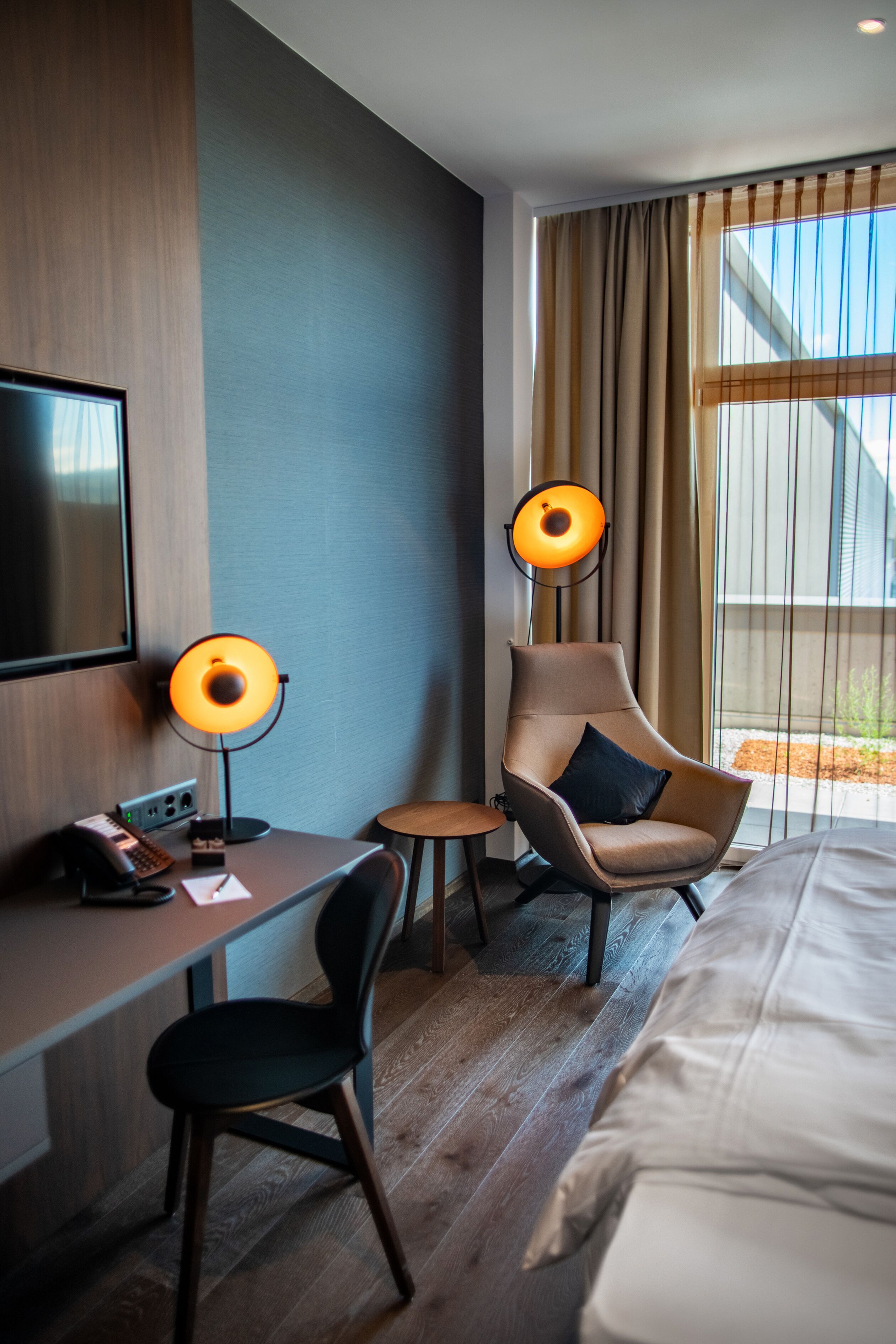 Habitación de hotel con cama, escritorio con teléfono, dos sillones, lámpara y televisor.
