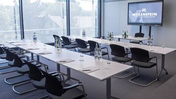 Un salón de conferencias con una pantalla grande mostrando 'MEILENSTEIN' y vistas de árboles al exterior.