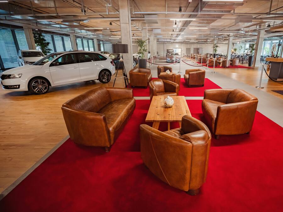 A modern showroom with a white car, leather chairs, a wooden table, and a red carpet.
