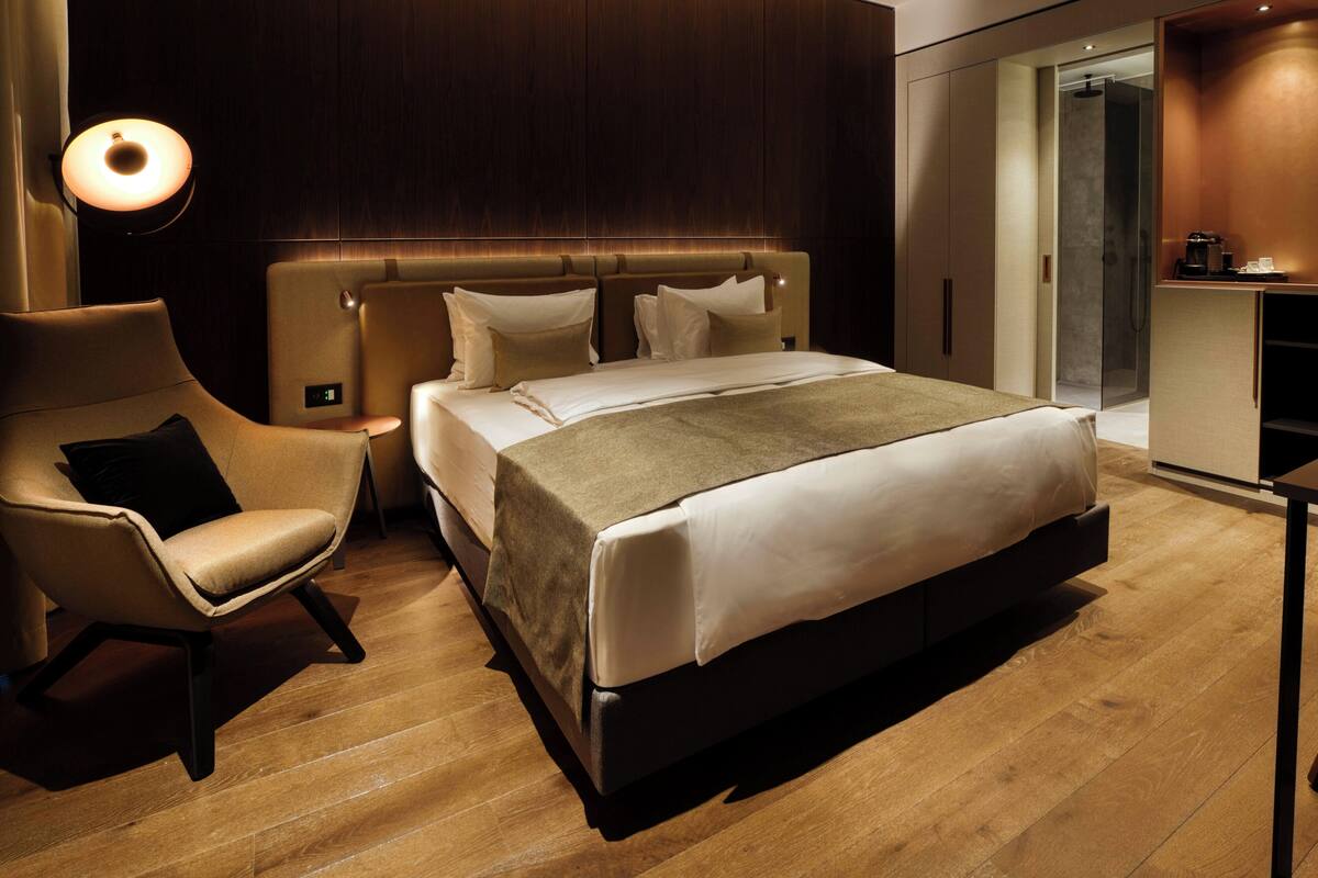 Una habitación de hotel moderna con una cama grande, un espacio de descanso con una silla, una mesita de noche y un armario empotrado.