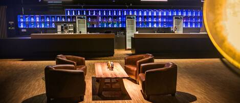 Una zona de bar con una mesa de madera y sillas de cuero, un cantinero y una exhibición iluminada en azul de botellas.