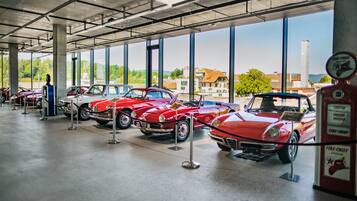 A museum with vintage cars on display, large windows, and a view of buildings and trees outside.