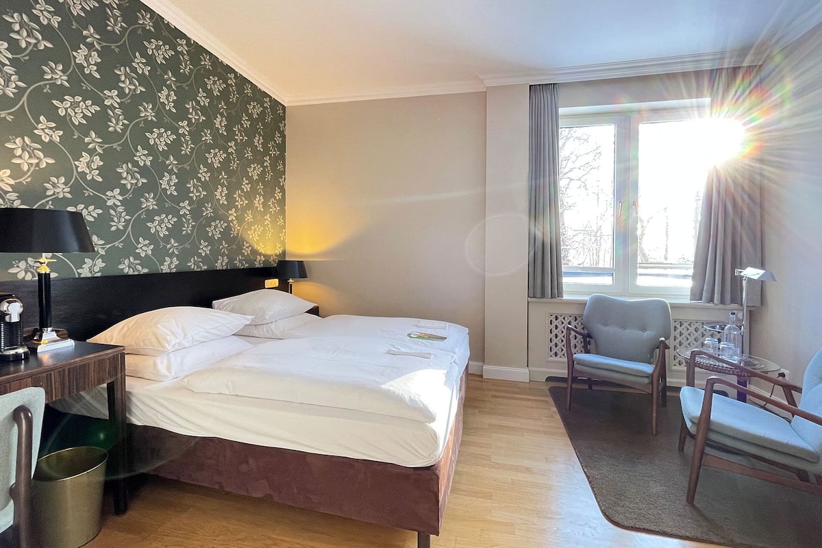 Hotellihuone, jossa on sänky, työpöytä lampun kanssa, tuoli ja ikkuna, jossa on verhot.