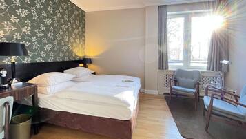 Una habitación de hotel con una cama, un escritorio con lámpara, una silla y un ventanal con cortinas.