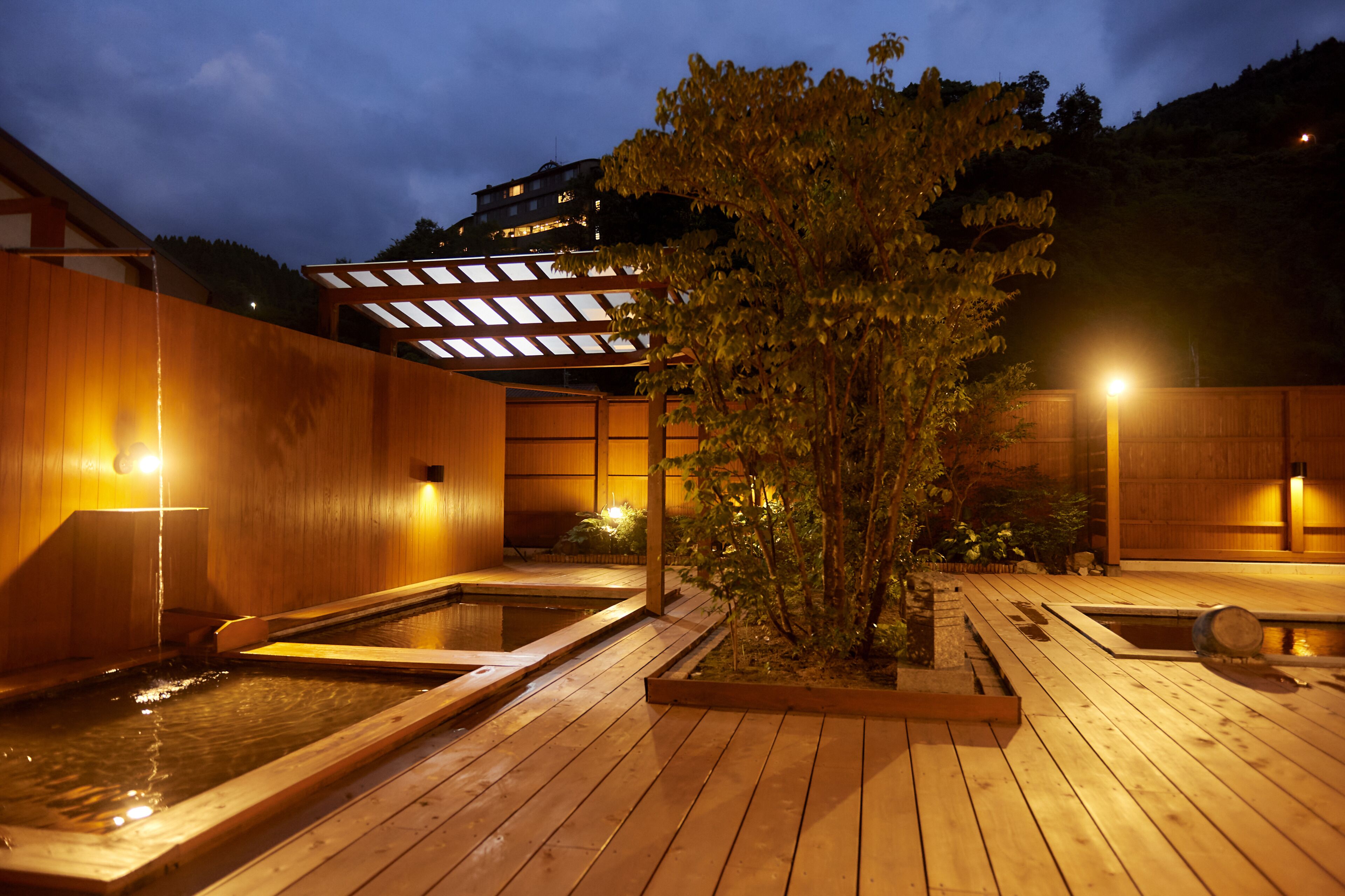 A wooden deck with hot tubs and a tree, surrounded by wooden walls and a building in the background.