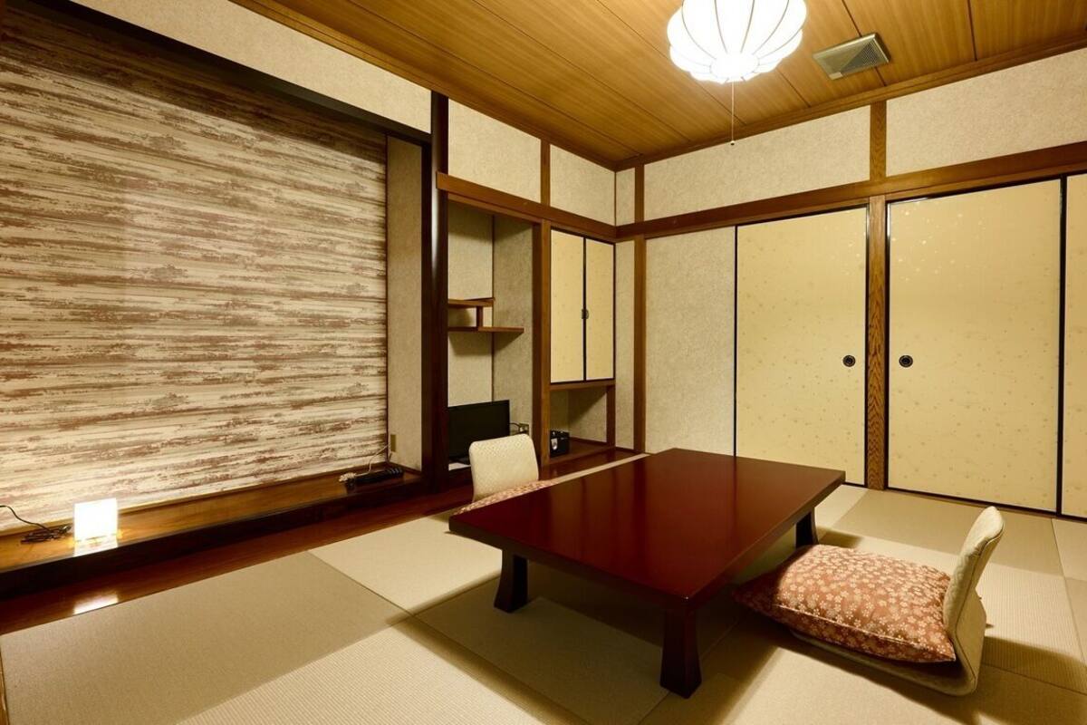 Ein traditioneller japanischer Raum mit Tatami-Boden, einem niedrigen Holztisch, einer gepolsterten Sitzbank, Schiebetüren und einer Deckenleuchte.