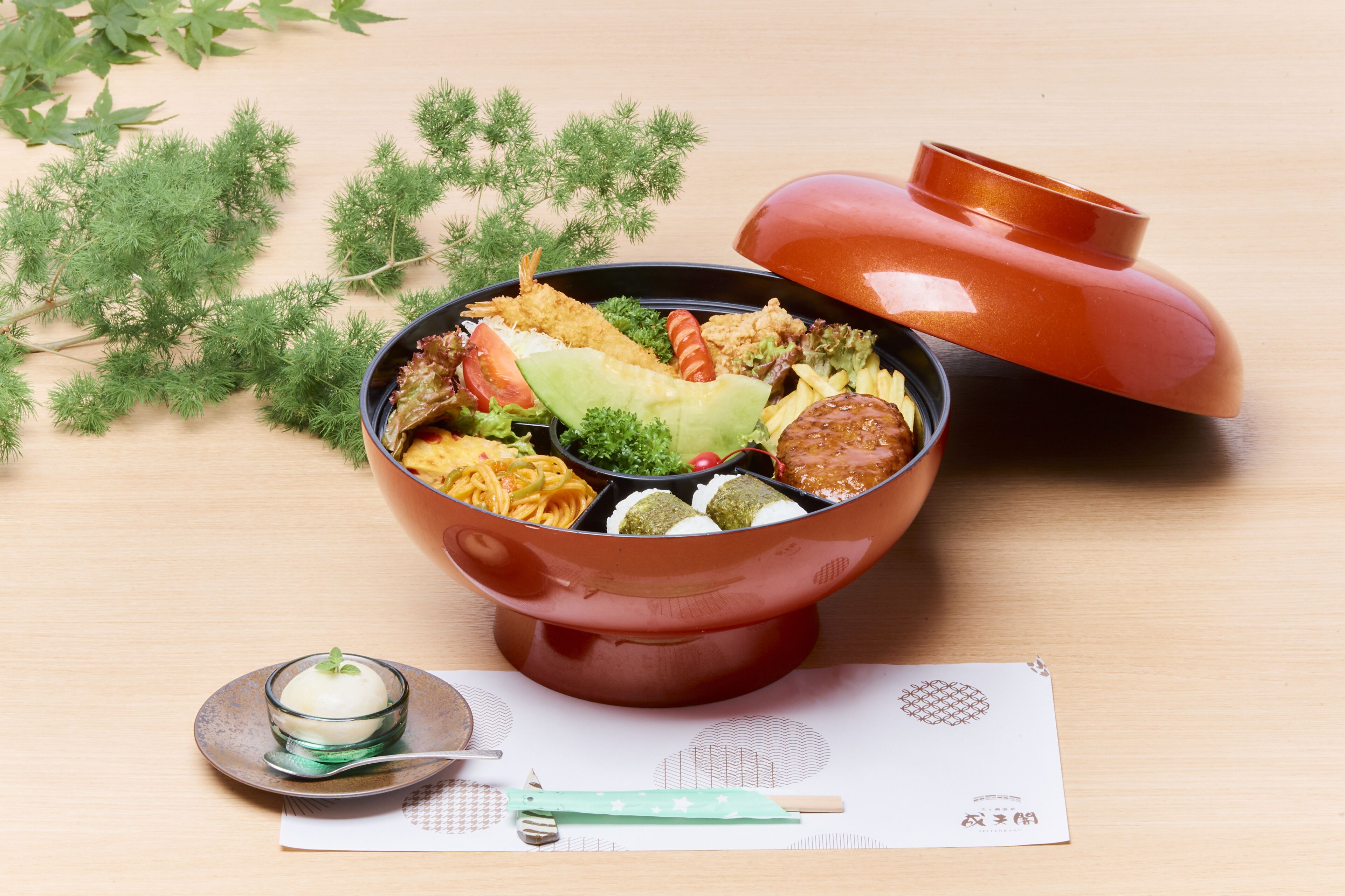 A bowl of food with a lid, a small bowl of soup, and a spoon on a table with a placemat.