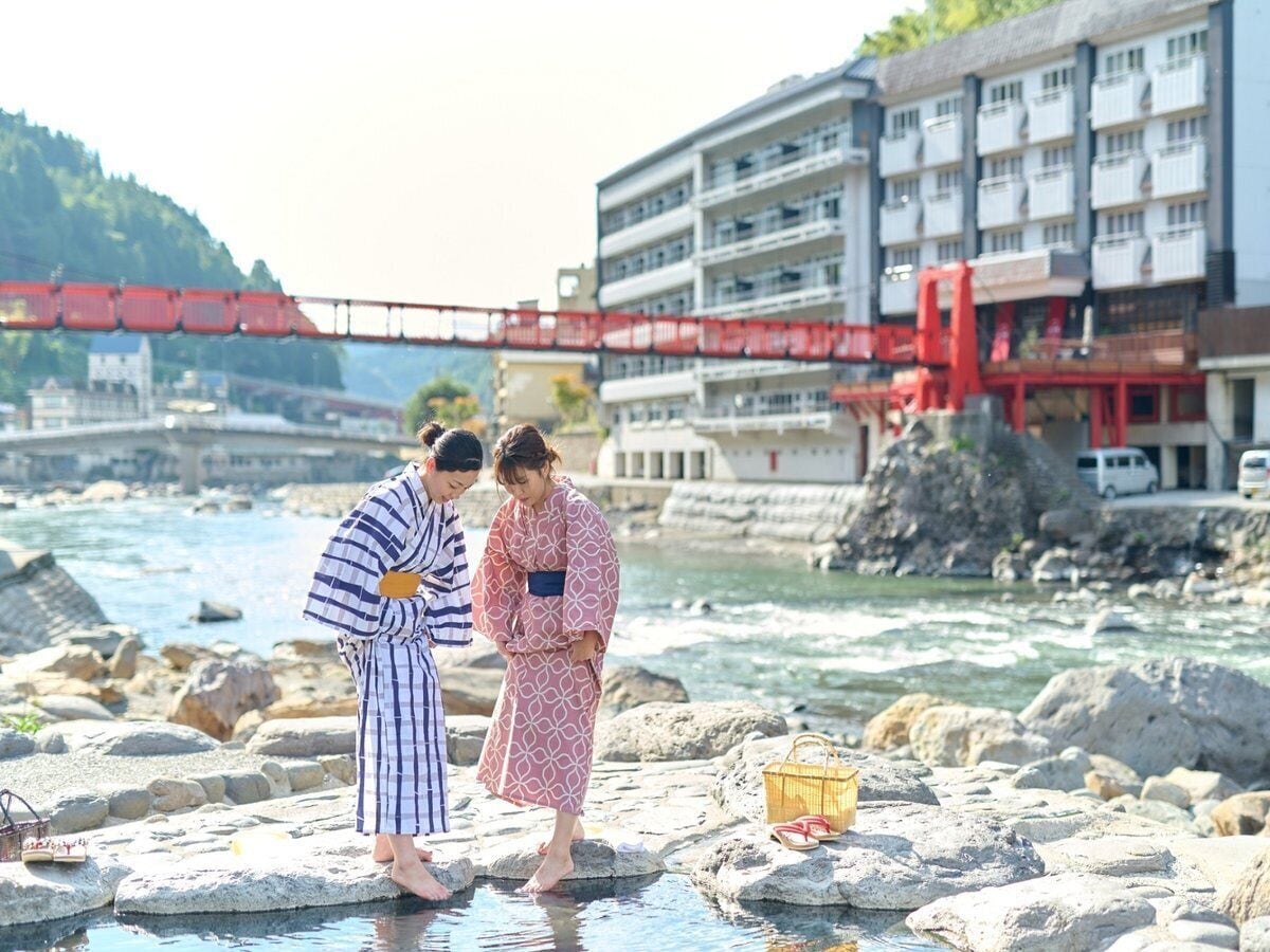 Two people standing on rocks by a river with a bridge in the background.