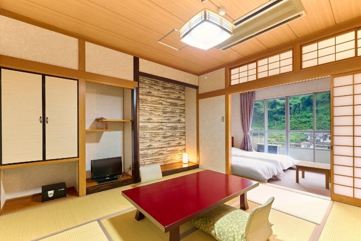 Ein traditioneller japanischer Raum mit Tatami-Böden, einem niedrigen Tisch, einem Fernseher, einem Bett und Blick ins Grüne.