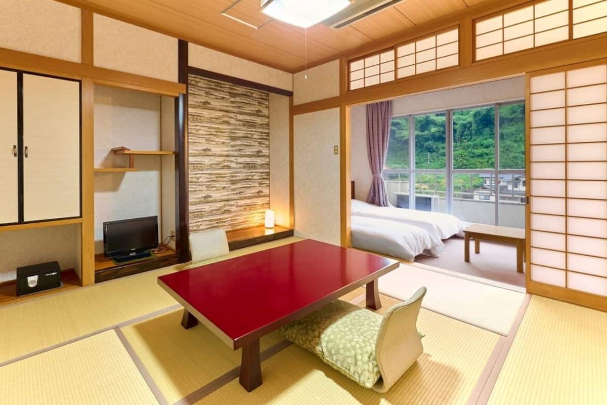 Ein traditioneller japanischer Raum mit Tatami-Böden, einem niedrigen Tisch, einem Fernseher und einem Bett mit Blick auf Grünflächen.