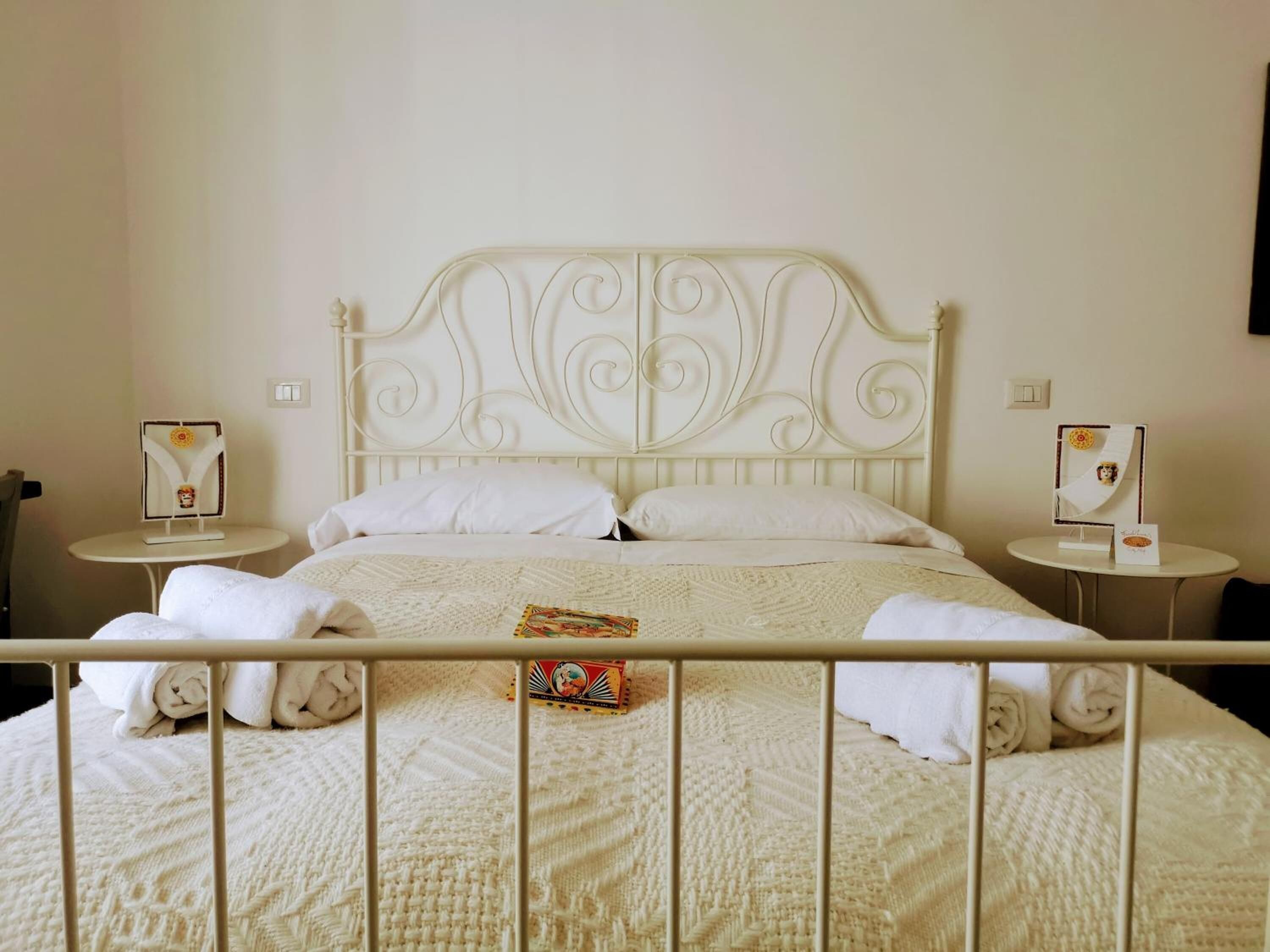 Een bed met een wit metalen hoofdeinde, twee nachtkastjes met decoratieve objecten, en een witte spreidek.