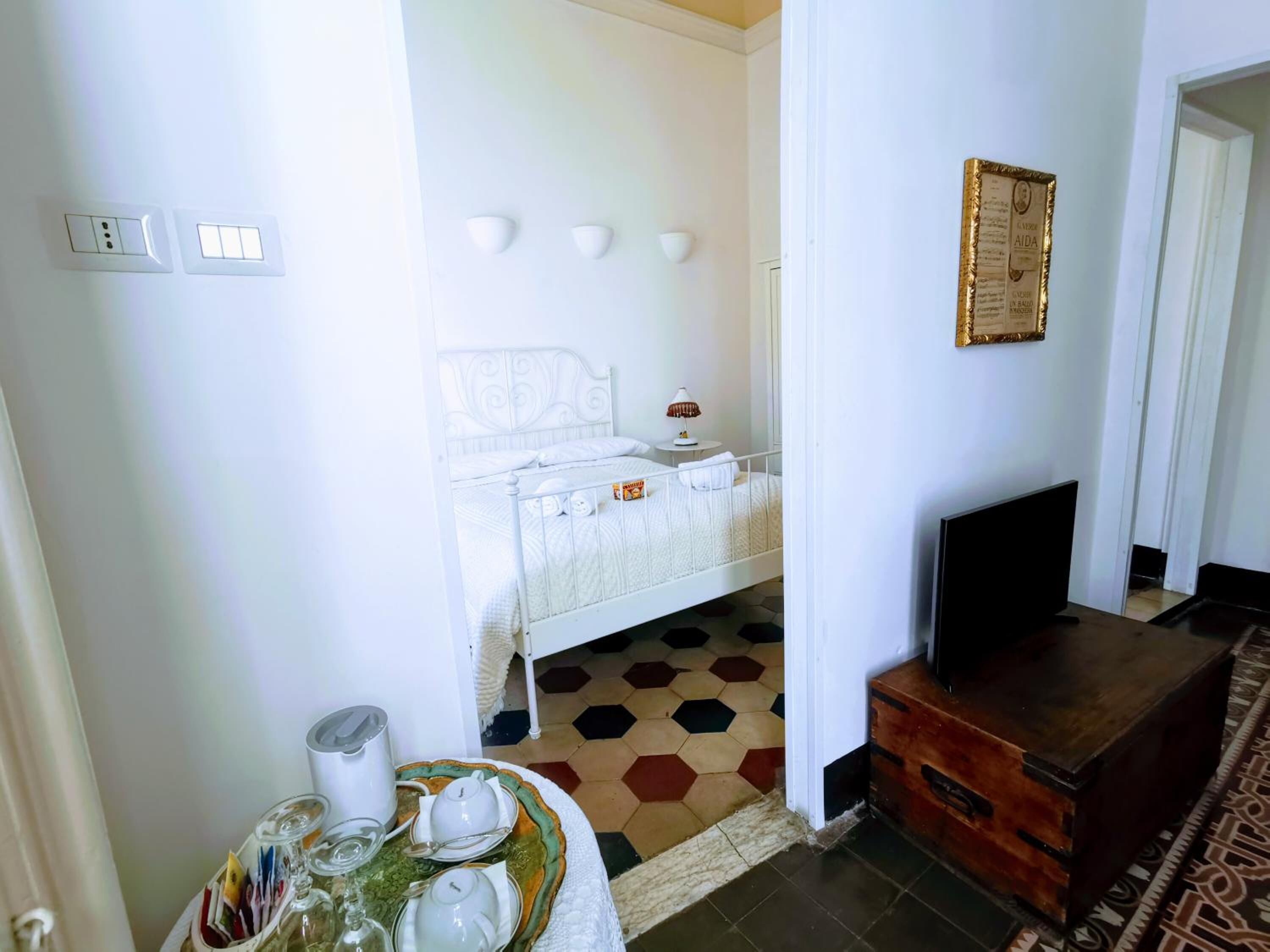 A hotel room with a bed, bedside table, lamp, TV, and a wooden chest of drawers.