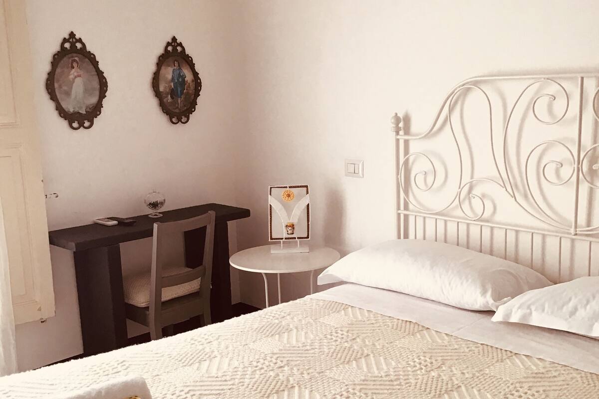 Ein Schlafzimmer mit einem Bett, Kissen, einem Nachttisch, einem Sessel und zwei gerahmten Bildern an der Wand.