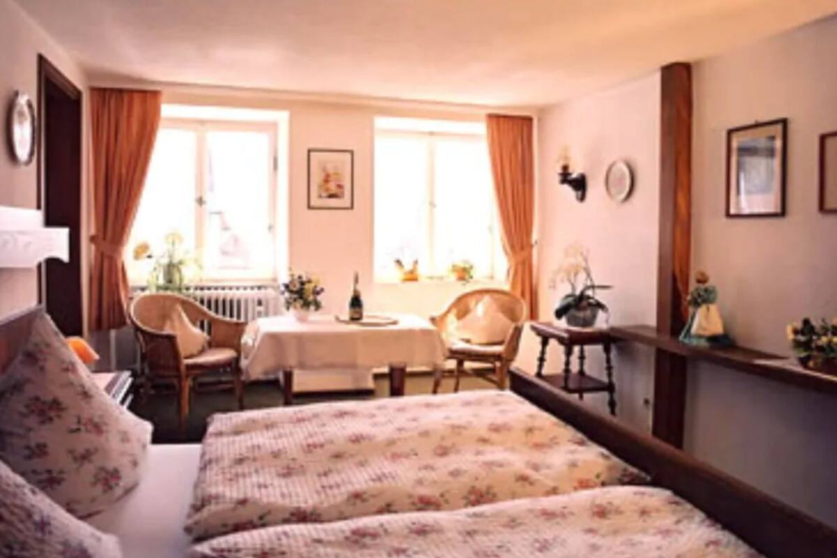 A cozy room with a bed, a table, and two chairs. There are windows with curtains, a radiator, and a clock on the wall.